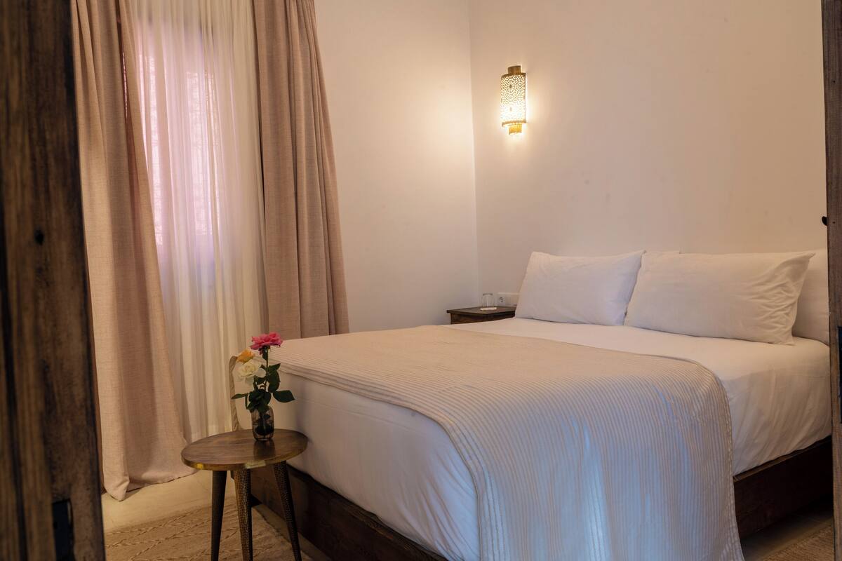 A neatly made bed with white linens, a bedside table with a vase of flowers, and a wall-mounted light fixture.