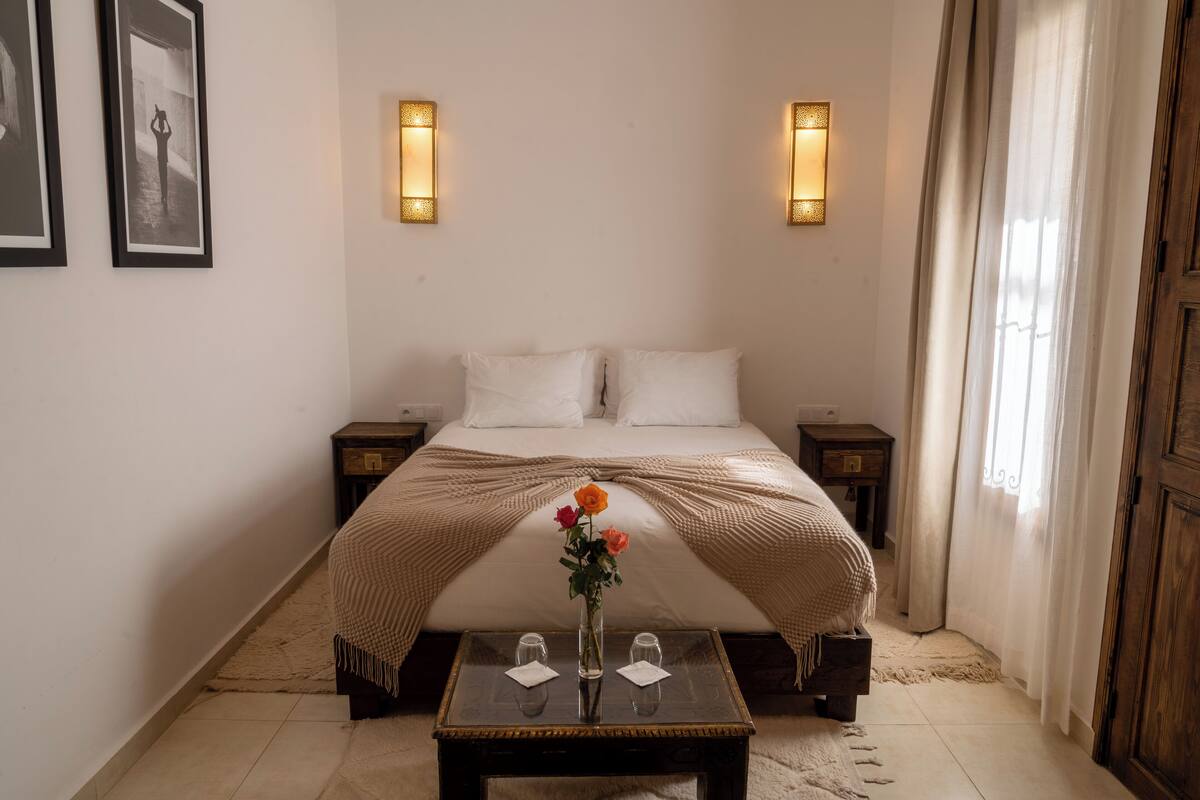 A neatly arranged hotel room with a bed, bedside tables, a small table with a vase of flowers, and two wall-mounted lights.
