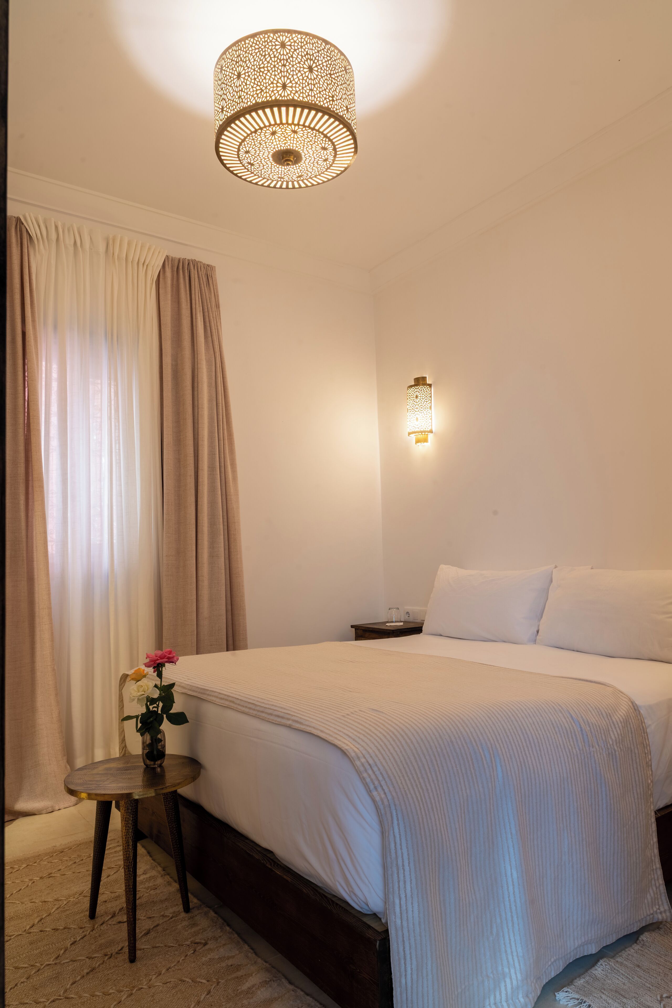 Ein Schlafzimmer mit einem Bett, einem Nachttisch, einer Wandlampe, einem Fenster mit Vorhängen und einem kleinen Tisch mit Blumen.
