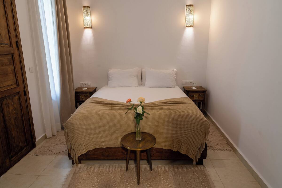 A bedroom with a bed, two bedside tables, a small round table with a vase of flowers, and two wall-mounted lamps.