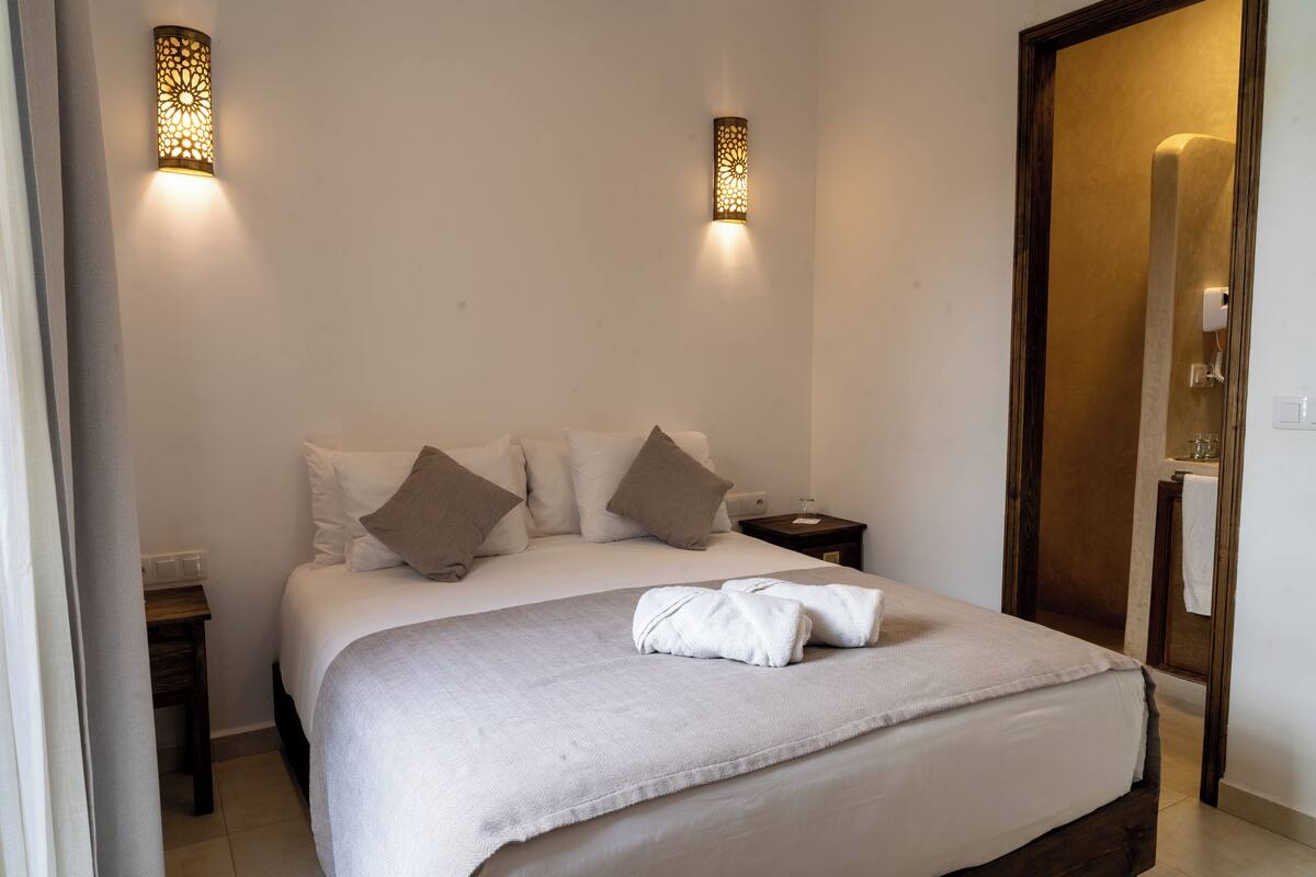 A neatly made bed with a white comforter and two pillows, a bedside table with a lamp, and a wall-mounted light fixture.