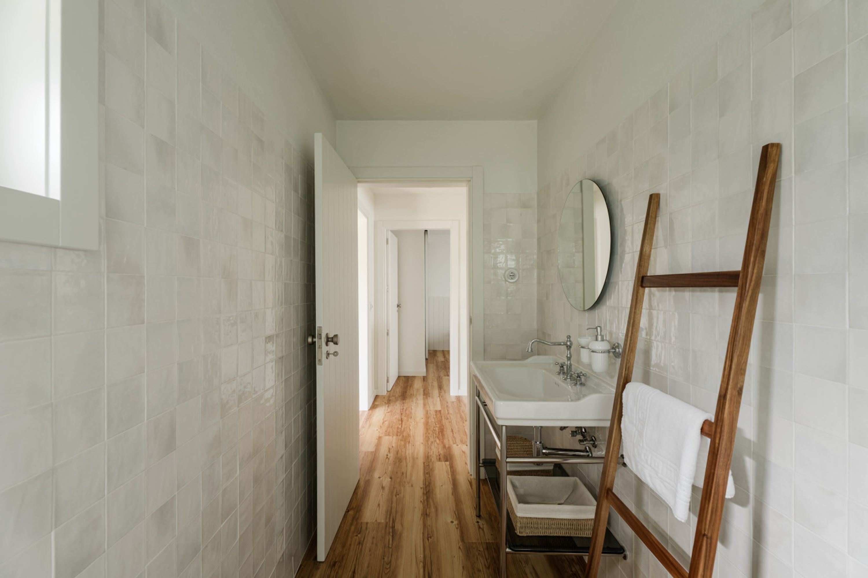 A narrow hallway with a wooden floor leading to a bathroom with a sink and mirror.