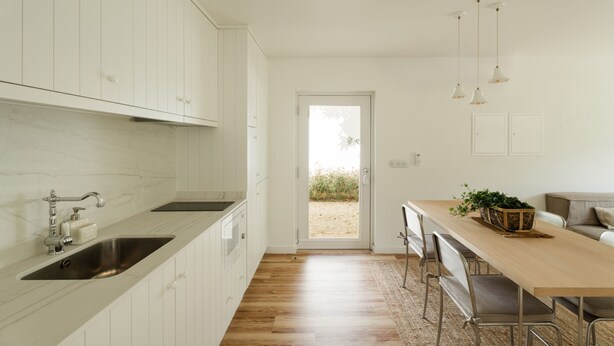 Una cocina moderna con armarios blancos, encimera de madera y un lavamanos empotrado. Hay una zona de comedor con una mesa larga y sillas, y vistas al exterior a través de una puerta.