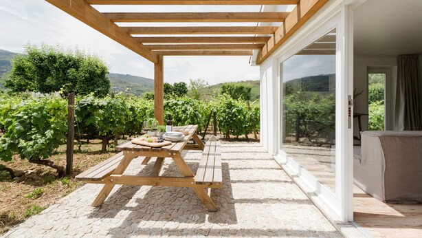 A covered patio with wooden benches, a table set with plates and wine glasses, and a view of a vineyard. - Imago Houses 3 - by MET (Lamego)