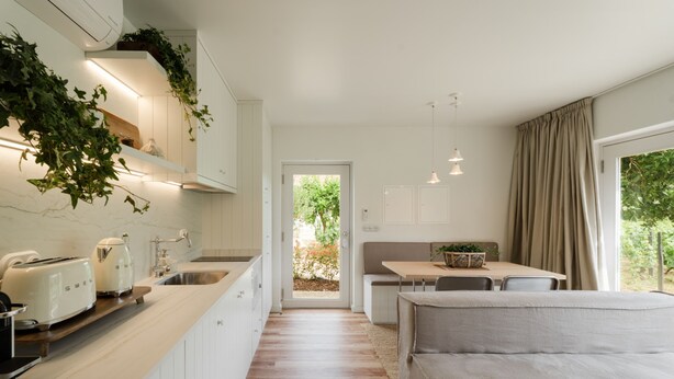 Una cocina moderna con armarios blancos, encimera de madera y horno empotrado. Cuenta con un comedor con banco y mesa, y un ventanal con cortinas.