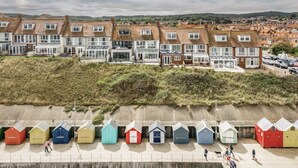 Una hilera de coloridas cabañas de playa a lo largo de un paseo marítimo, con casas al fondo.