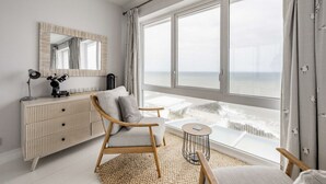 Un dormitorio moderno con un ventanal grande, una silla de madera, una mesita de noche y un aparador blanco.