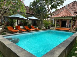 A swimming pool with lounge chairs and umbrellas, surrounded by greenery and a building with a tiled roof. - Villa Tiga Naga Sanur (Denpasar)