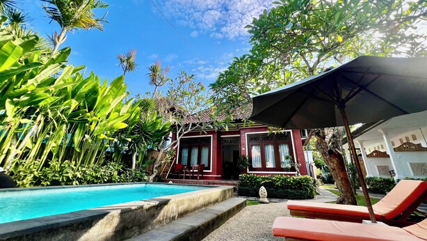 A pool area with lounge chairs and an umbrella, surrounded by lush greenery and a building with a red facade. - Villa Tiga Naga Sanur (Denpasar)