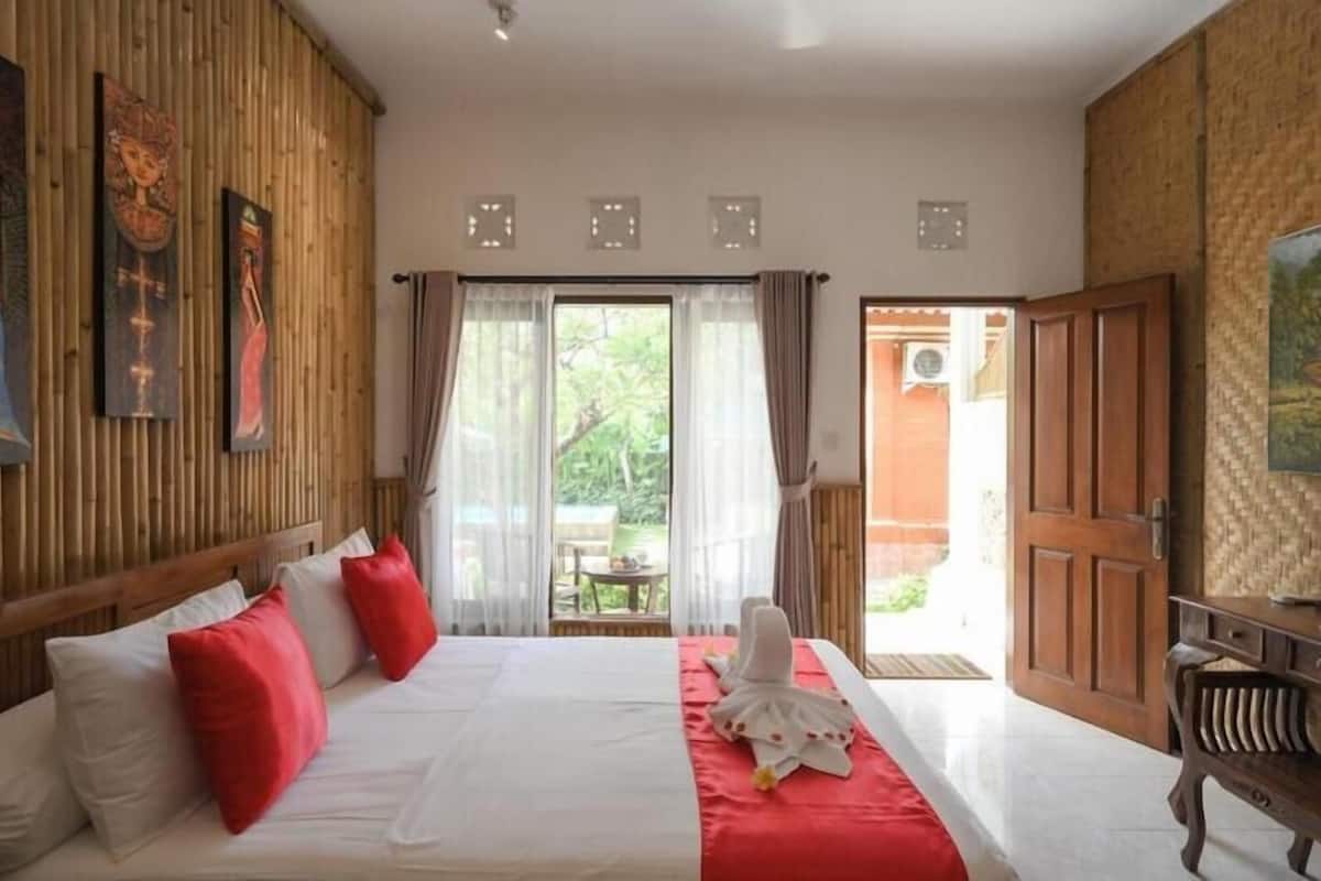 Une chambre d’hôtel avec un lit, une tête de lit en bois, une couvre-lit rouge et blanche, et une vue sur l’extérieur.