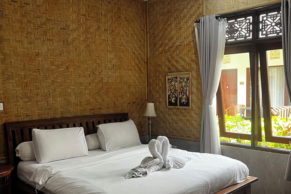 Une chambre d’hôtel avec une tête de lit en bois, un lit avec du linge de maison blanc, un ventilateur de plafond et une fenêtre avec des rideaux.