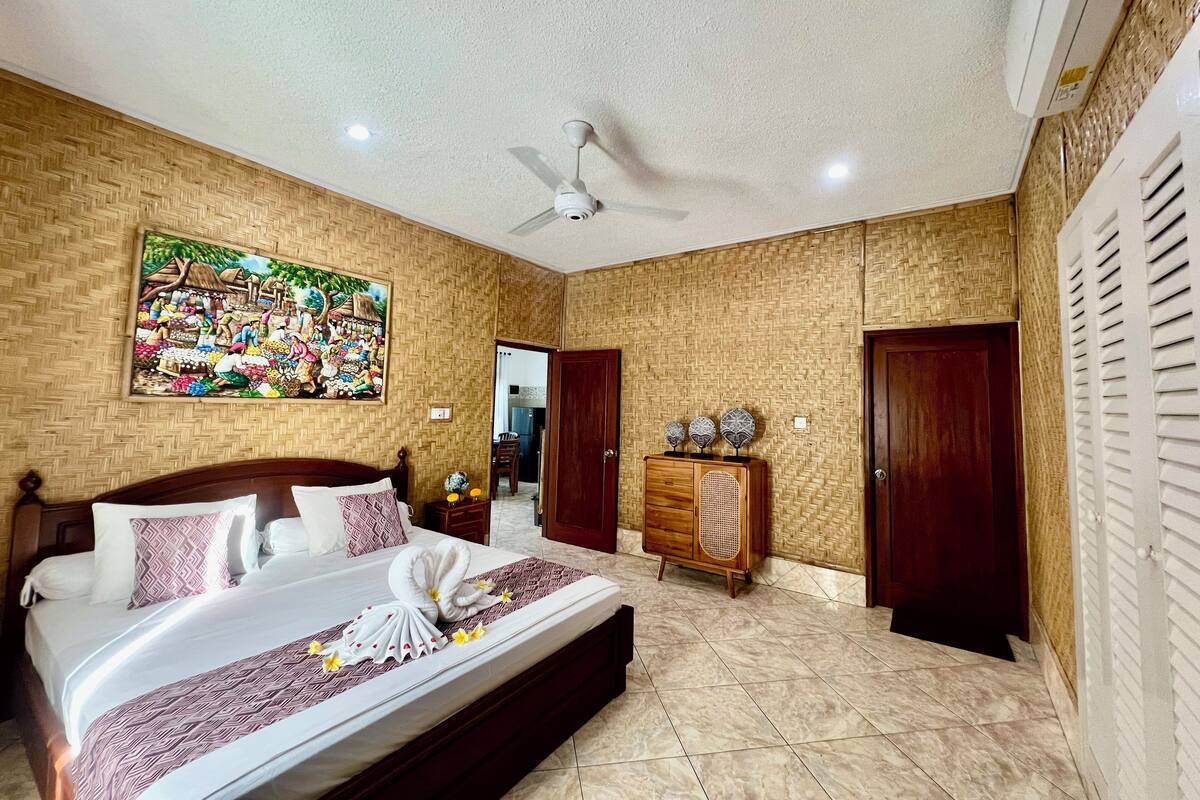 Une chambre d’hôtel dotée d’un grand lit, d’un ventilateur de plafond, d’une tête de lit en bois, d’un climatiseur mural, d’une armoire en bois agrémentée d’objets décoratifs et d’un mur orné d’un motif en chevrons.