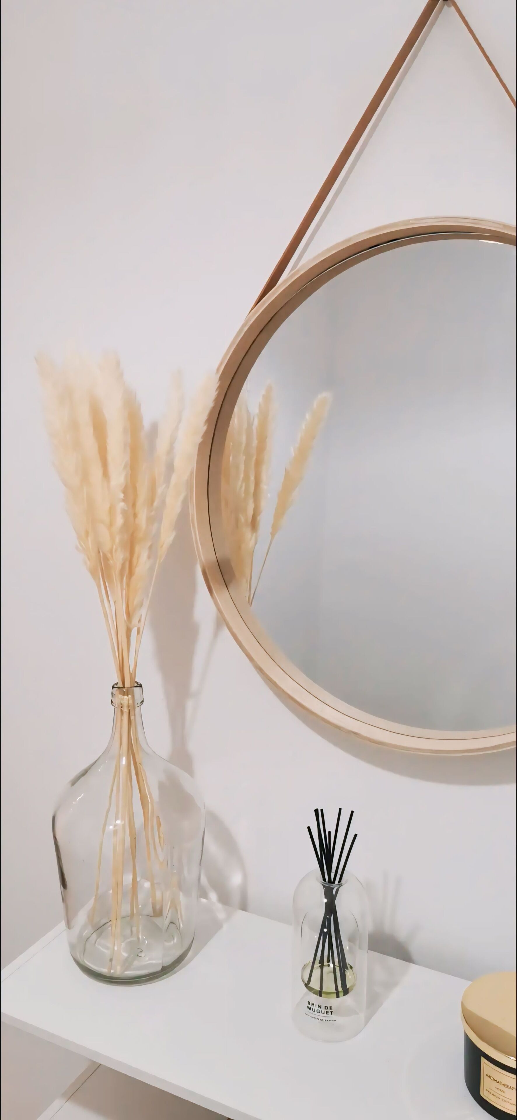 Een spiegel met houten lijst, een glazen vaas met pampa gras, en een glazen pot met rietstengels op een witte ondergrond.