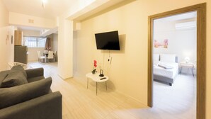 Ein modernes Hotelzimmer mit einem an der Wand befestigten Flachbildfernseher, einem kleinen weißen Beistelltisch mit Blumenvase und einem Bett mit weißem Kopfteil.