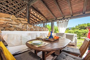 A covered outdoor seating area with stone walls, a wooden table with plates and fruit, and a view of greenery. - Il Vecchio Faro Lilli (Loiri Porto San Paolo)