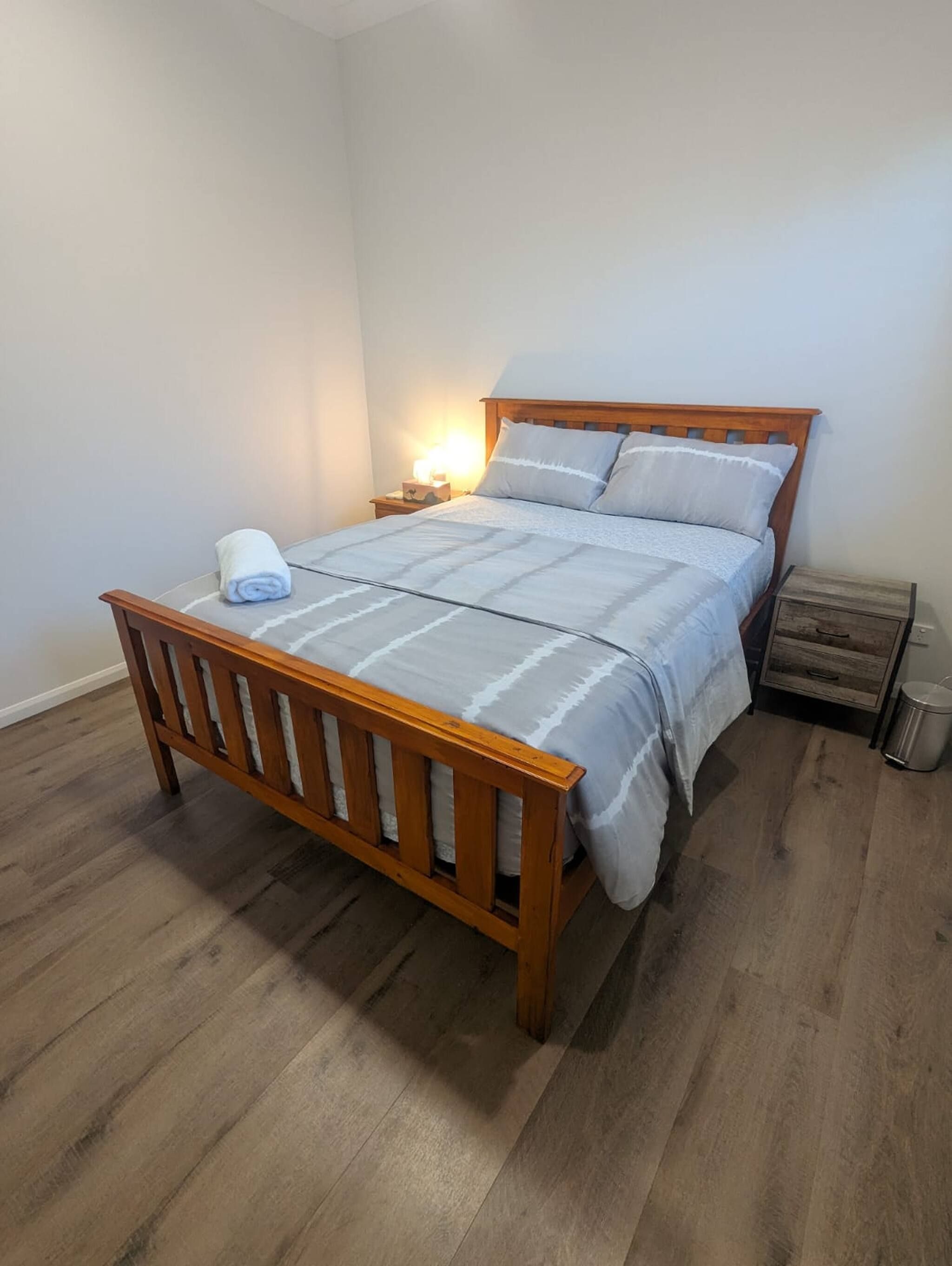 A single bed with a wooden frame and a white bedspread, a bedside table with a lamp, and a trash bin.