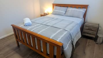 A single bed with a wooden frame and a white bedspread, a bedside table with a lamp, and a trash bin.