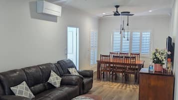 A spacious living room with a leather sofa, a wooden coffee table, and a dining area with a wooden table and chairs. There are ceiling fans and an air conditioning unit.