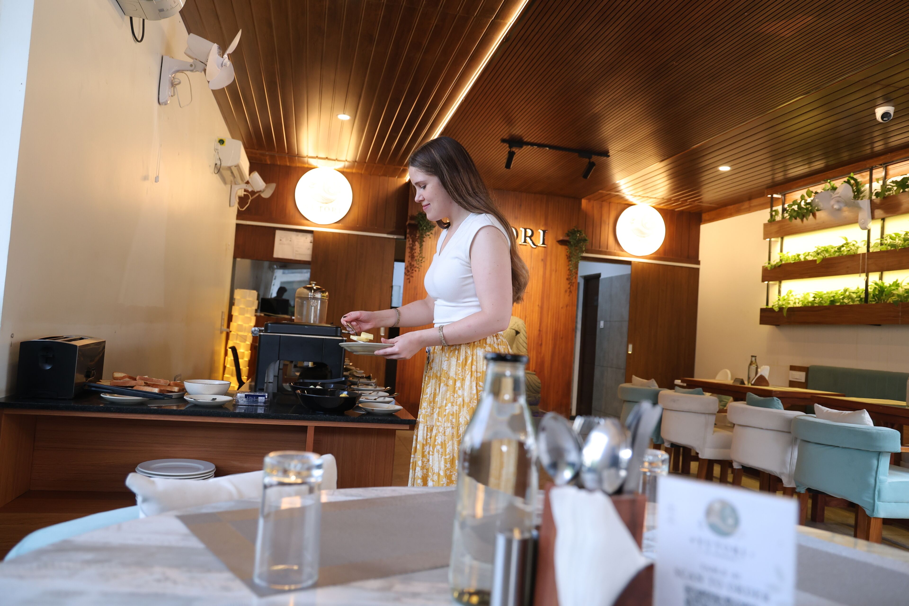 A woman in a white top and yellow skirt standing at a counter with a coffee machine, plates, and a menu in a restaurant with wooden ceiling, plants, and seating area.