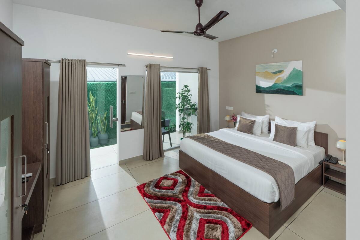 Une chambre moderne avec un grand lit, un ventilateur de plafond, une table de chevet avec une lampe et une vue sur un espace extérieur verdoyant.