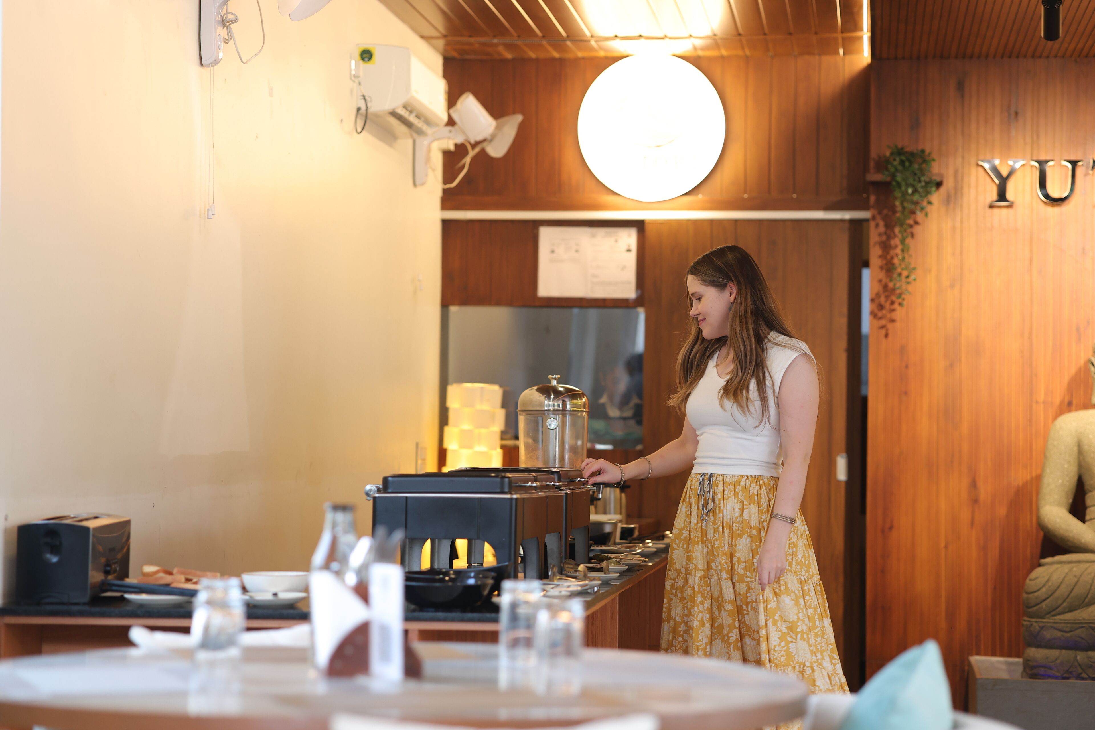 Una mujer con una blusa blanca y falda amarilla está de pie junto a una barra con una máquina de café, en una habitación con paredes de madera y un cartel que dice 'YU'.