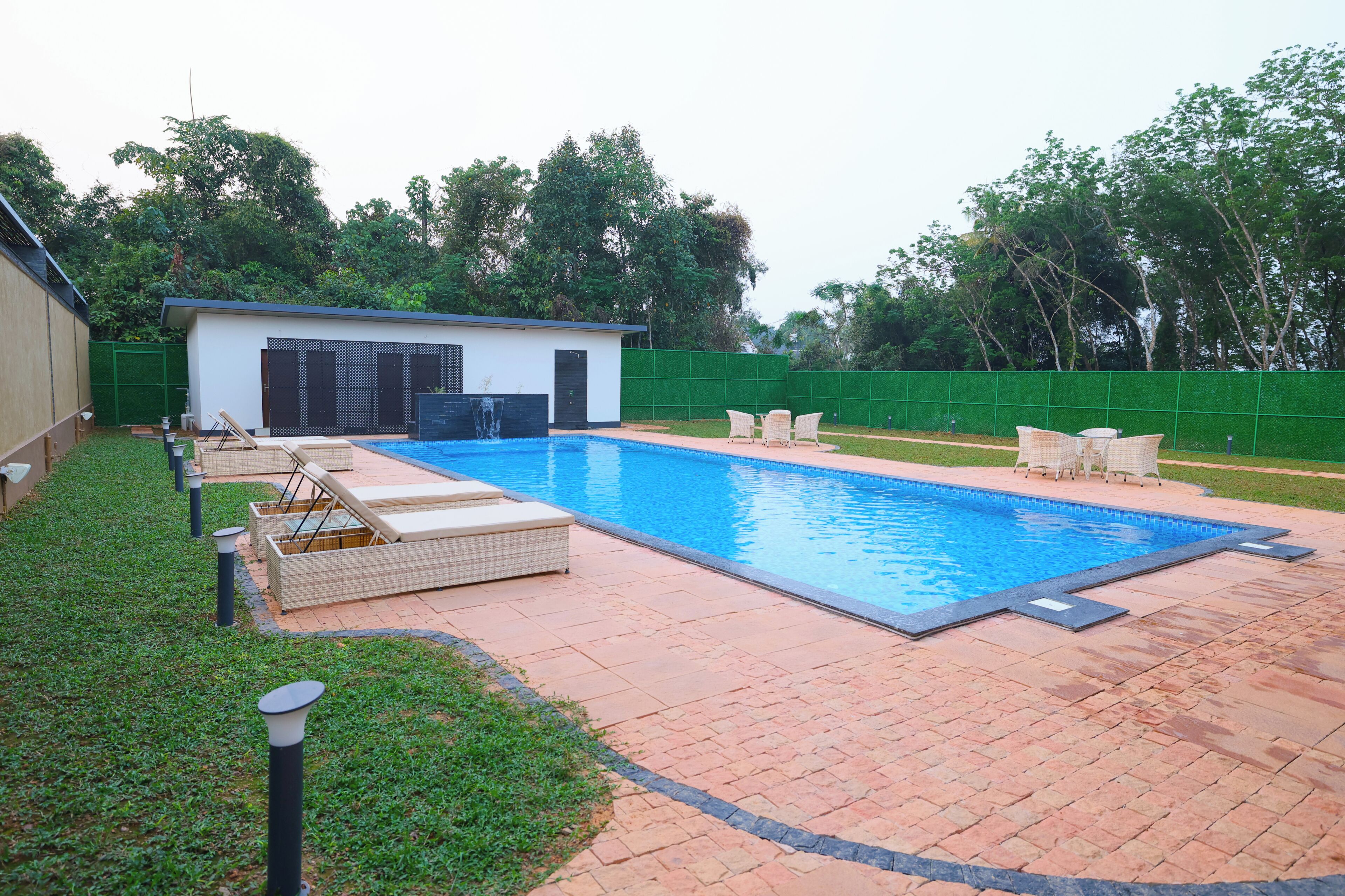 Une piscine rectangulaire avec une terrasse carrelée, un petit bâtiment blanc doté d’une porte sombre et une clôture verte.