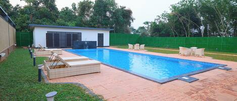 Une piscine rectangulaire avec une terrasse carrelée, un petit bâtiment blanc doté d’une porte sombre et une clôture verte.