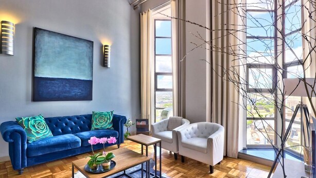 A modern living room with a blue sofa, a white armchair, and a coffee table. There is a large abstract painting on the wall and a window with a view of the city. - Cape Eazi Stayz Mutual Heights (Cape Town)