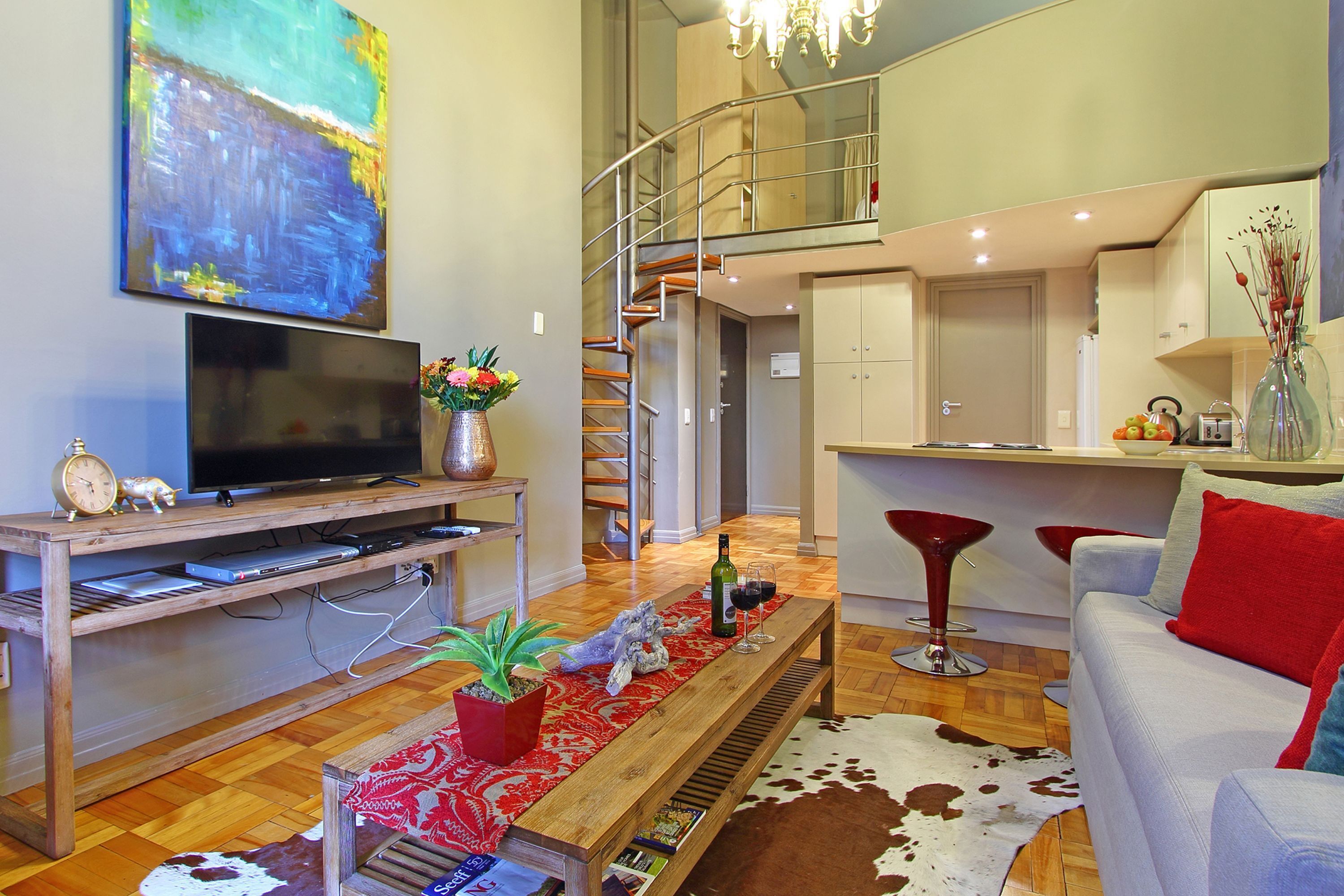 Un salon moderne avec un téléviseur, un meuble de rangement en bois, un tapis à motifs rouges et un tapis en peau de vache. Un escalier mène à un espace cuisine comprenant une table et des chaises.
