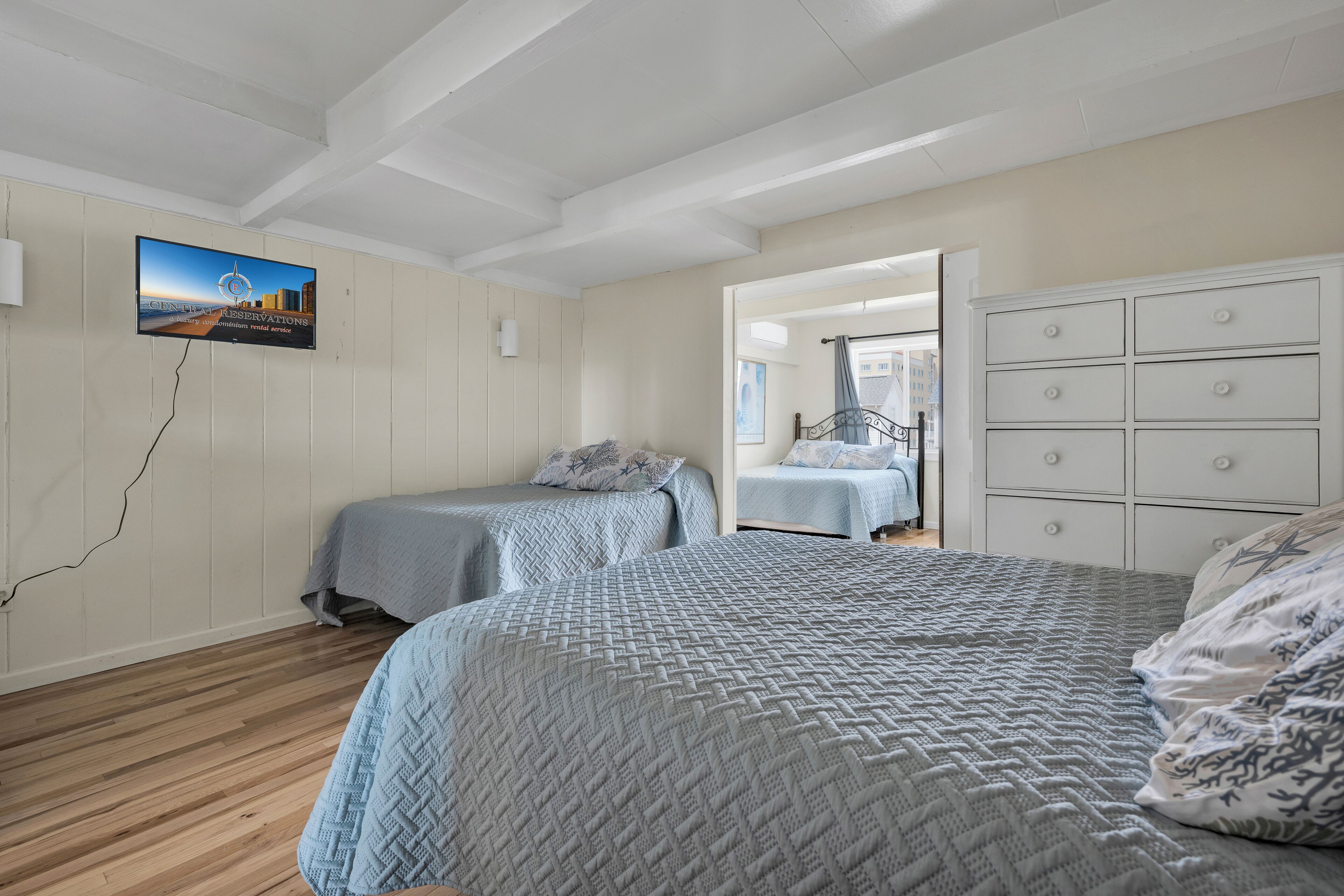 Quarto com duas camas, uma cômoda branca e televisão na parede.