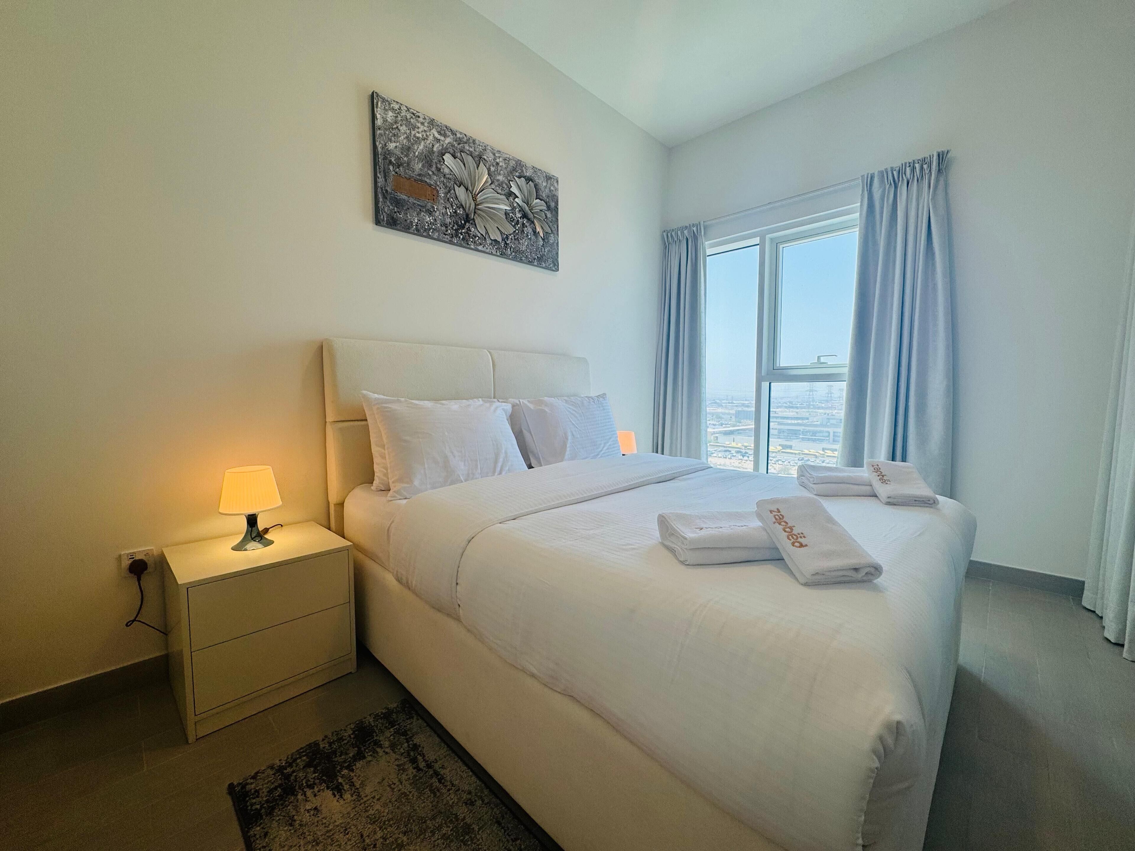 A hotel room with a large bed, a nightstand, a lamp, a window with curtains, and a city view.