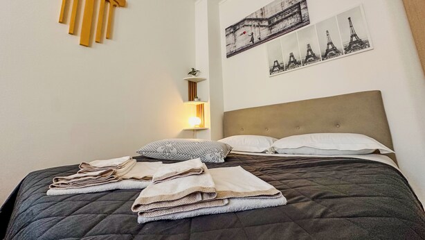 A neatly made bed with folded towels, a bedside lamp, and wall art depicting the Eiffel Tower. - Modern Studio Lingotto Historic District (Torino)