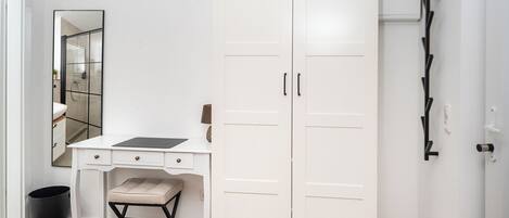 A room with a white wardrobe, a desk with a chair, a small stool, and a trash bin.