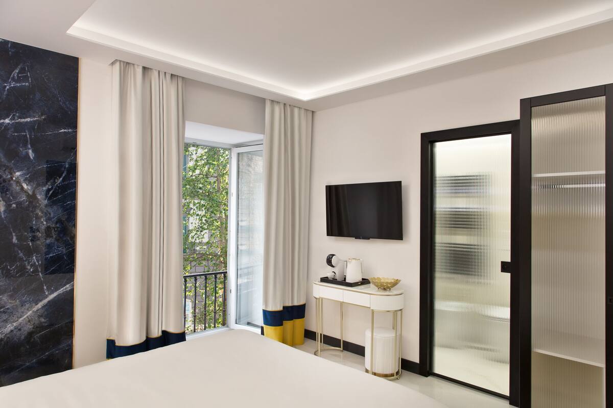 A modern hotel room with a large bed, a flat-screen TV mounted on the wall, a small table with a bowl and a lamp, and a sliding glass door leading to a balcony.