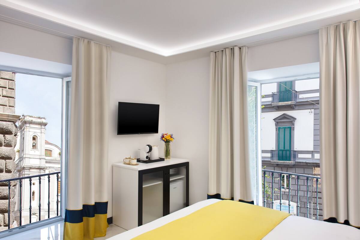 A hotel room with a bed, a TV mounted on the wall, a small desk with a vase of flowers, and a view of the cityscape through large windows.