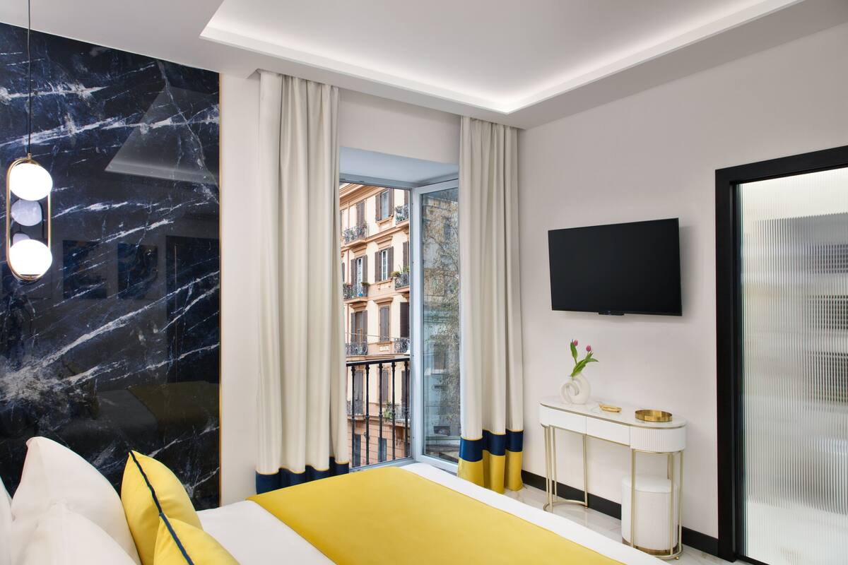A modern hotel room with a large bed, a bedside table, a TV, and a view of the city through a balcony.