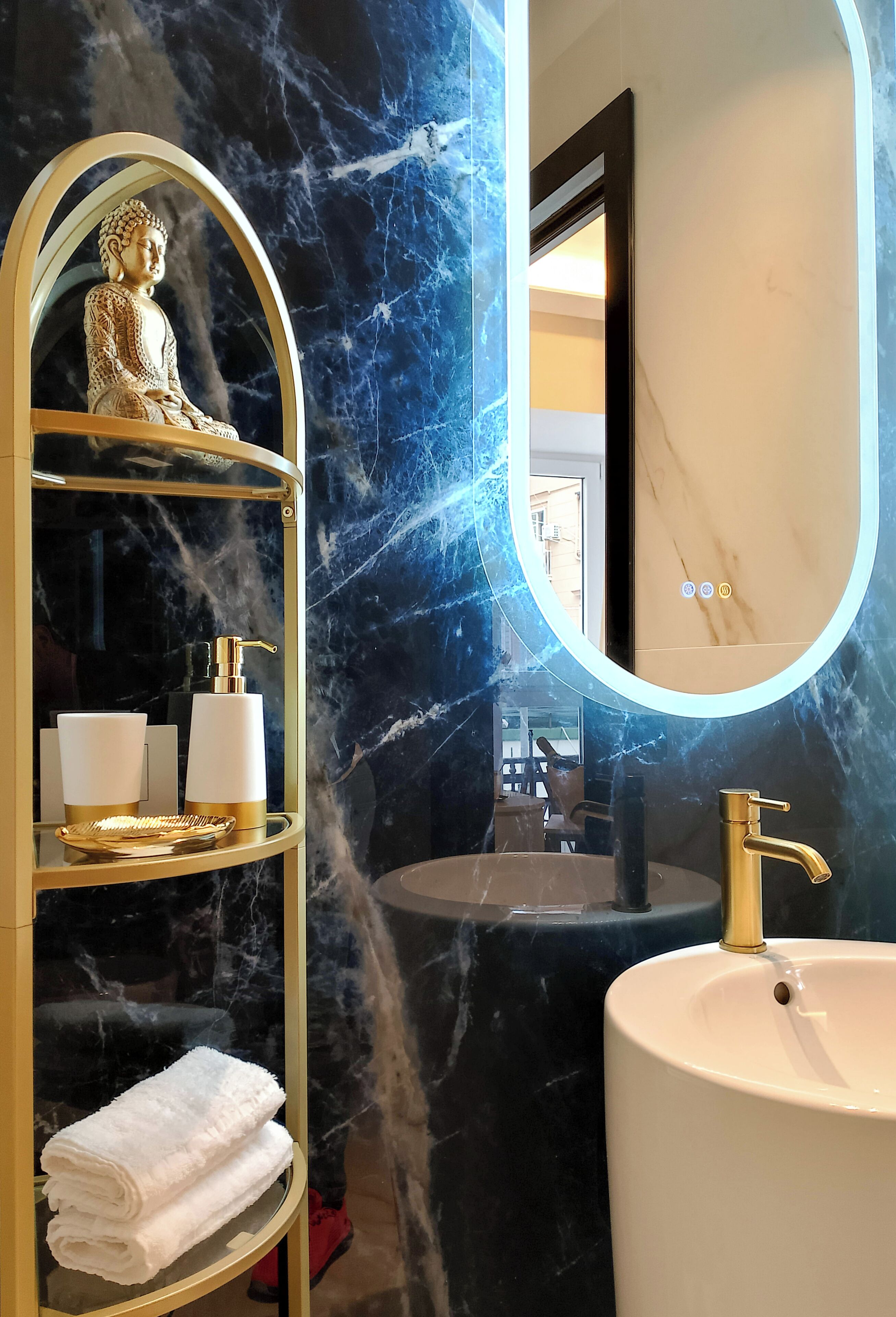 A bathroom with a marble wall, a gold-framed shelf with toiletries, a white sink, and a mirror.