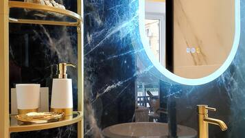 A bathroom with a marble wall, a gold-framed shelf with toiletries, a white sink, and a mirror.