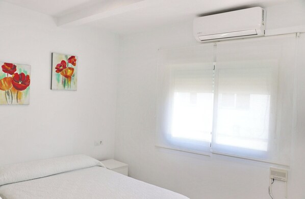 A white room with a bed, a window with blinds, and two paintings on the wall.