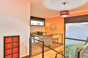 A modern interior with a large window offering a view of the sea, a red storage unit, a wicker chair, and a clock on the wall. - Villa Leonor Cliffside Retreat (Calheta)