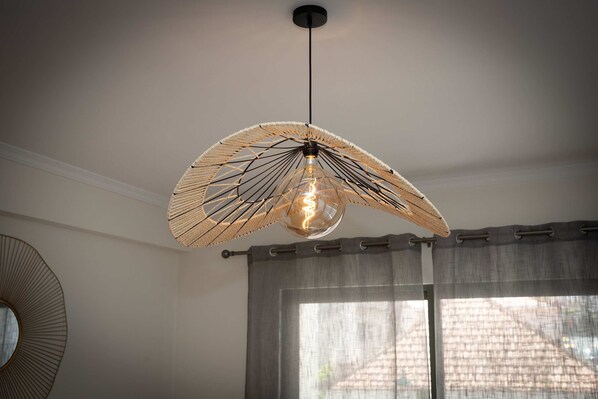 A modern pendant light with a unique fan-like design, hanging from a ceiling in a room with a large window and a round mirror on the wall. - Apartamento Janela do Atlantico (Machico)