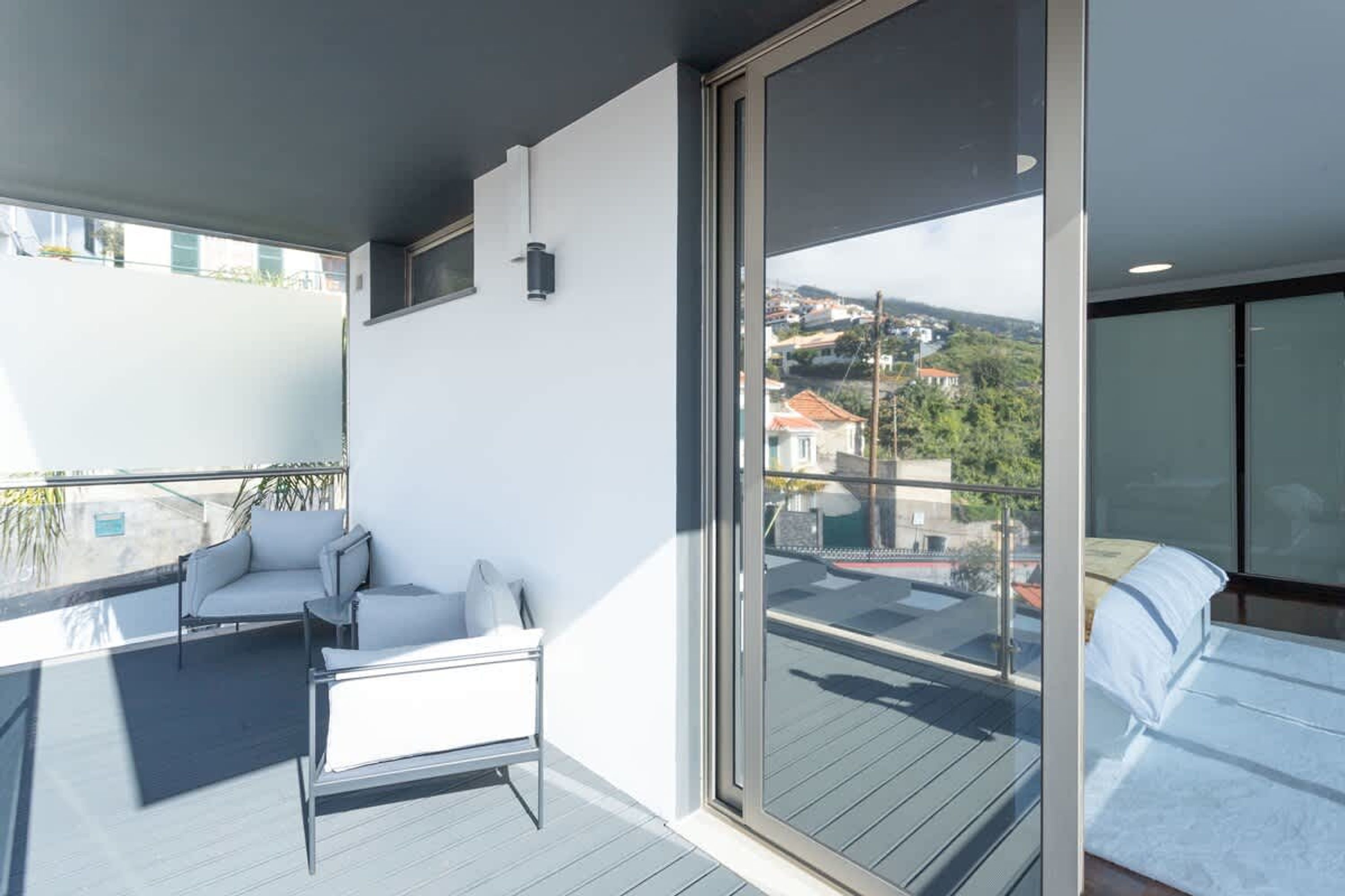 Ein Balkon mit einer Couch und einem Bett, ein großes Fenster und Blick auf Gebäude in der Ferne.