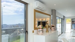 A modern interior with a large window, a white console table with decorative items, and a view of the cityscape. - Villa Andrade Ocean View (Funchal)