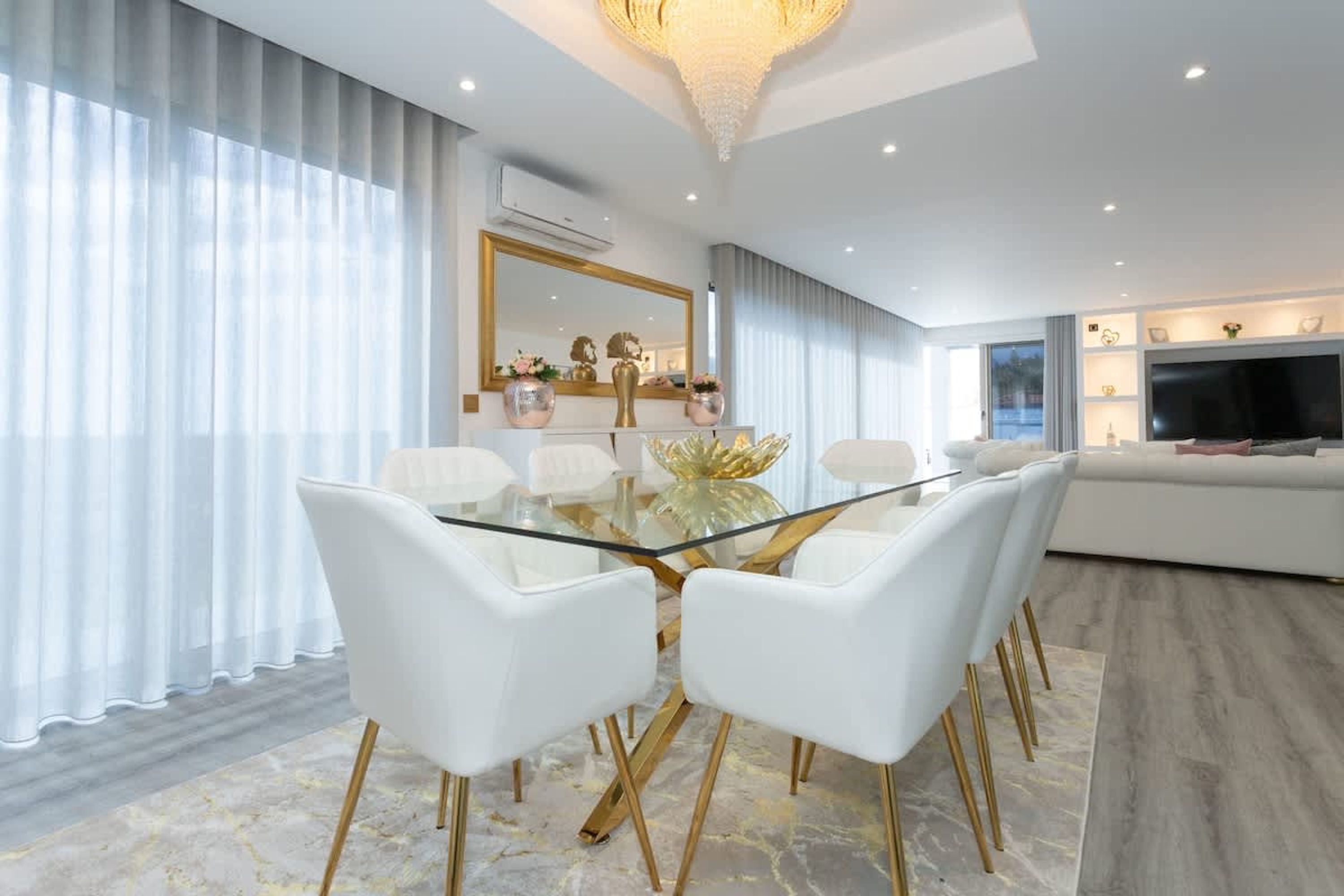 A modern dining area with a glass table and white chairs, a large mirror, and a television in the background.