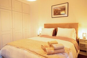 Een net opgemaakt bed met een geborduurd beddekleed en opgerolde handdoeken, een houten hoofdbord en een ingelijst foto aan de muur.