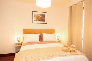 Een netjes opgemaakt bed met een bedspreide met patroon, twee bedlampen en een ingelijst foto aan de muur.