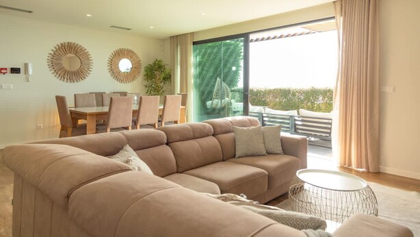 Una sala de estar moderna con un sofá seccional beige grande, un comedor con mesa y sillas de madera, y un balcón con vistas a zonas verdes.