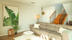 A living room with a beige sofa, a white coffee table, a large green plant, and a staircase with wooden steps. - Villa Eden Nature in Prazeres (Calheta)