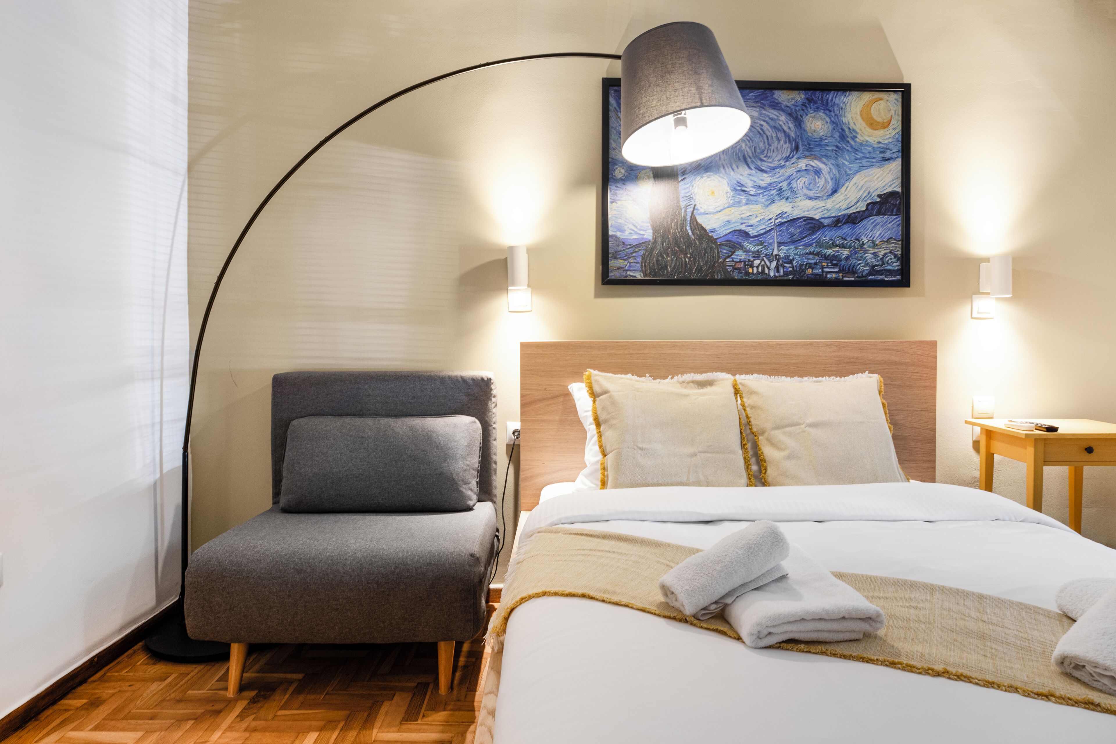 A hotel room with a bed, a chair, a lamp, and a painting on the wall.