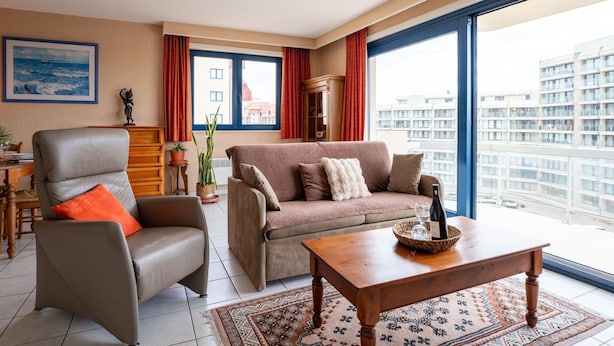 Un saló modern amb un sofà, una poltrona i una taula de centre. Hi ha una finestra gran amb vista a edificis i un quadre a la paret.